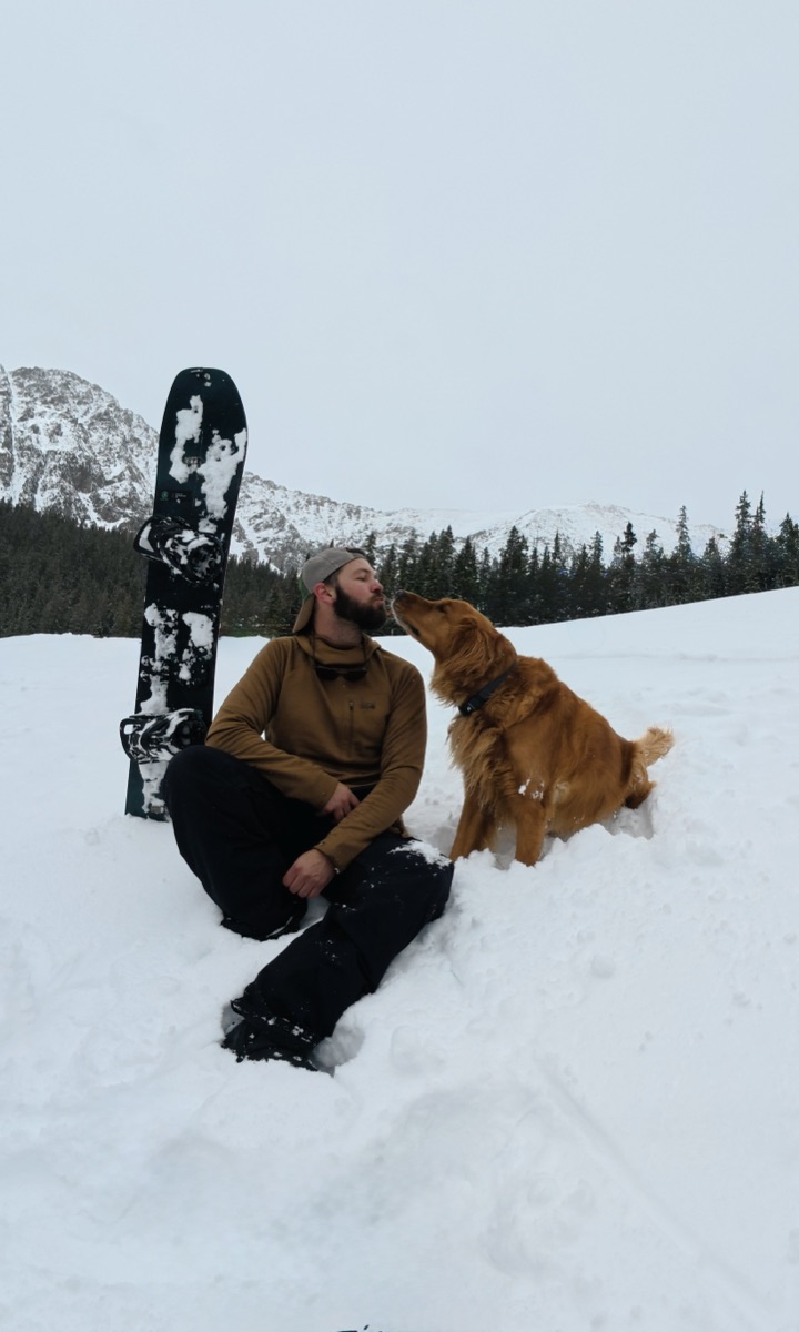 Christopher sitting in the snow beside his snowboard while Oliver leans in to lick his face