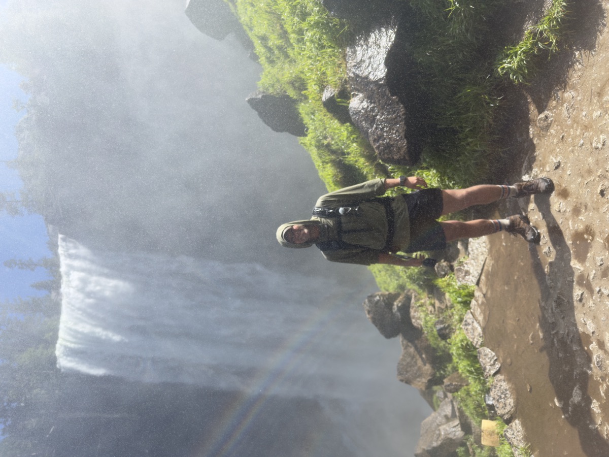 Christopher in a rain hood standing in the mist at the base of a tall waterfall